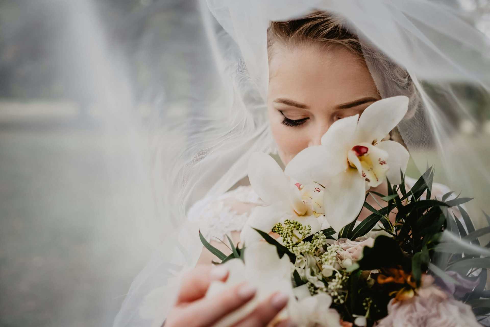 Bride with eyes closed, holding a bouquet of white orchids and greenery under a veil.